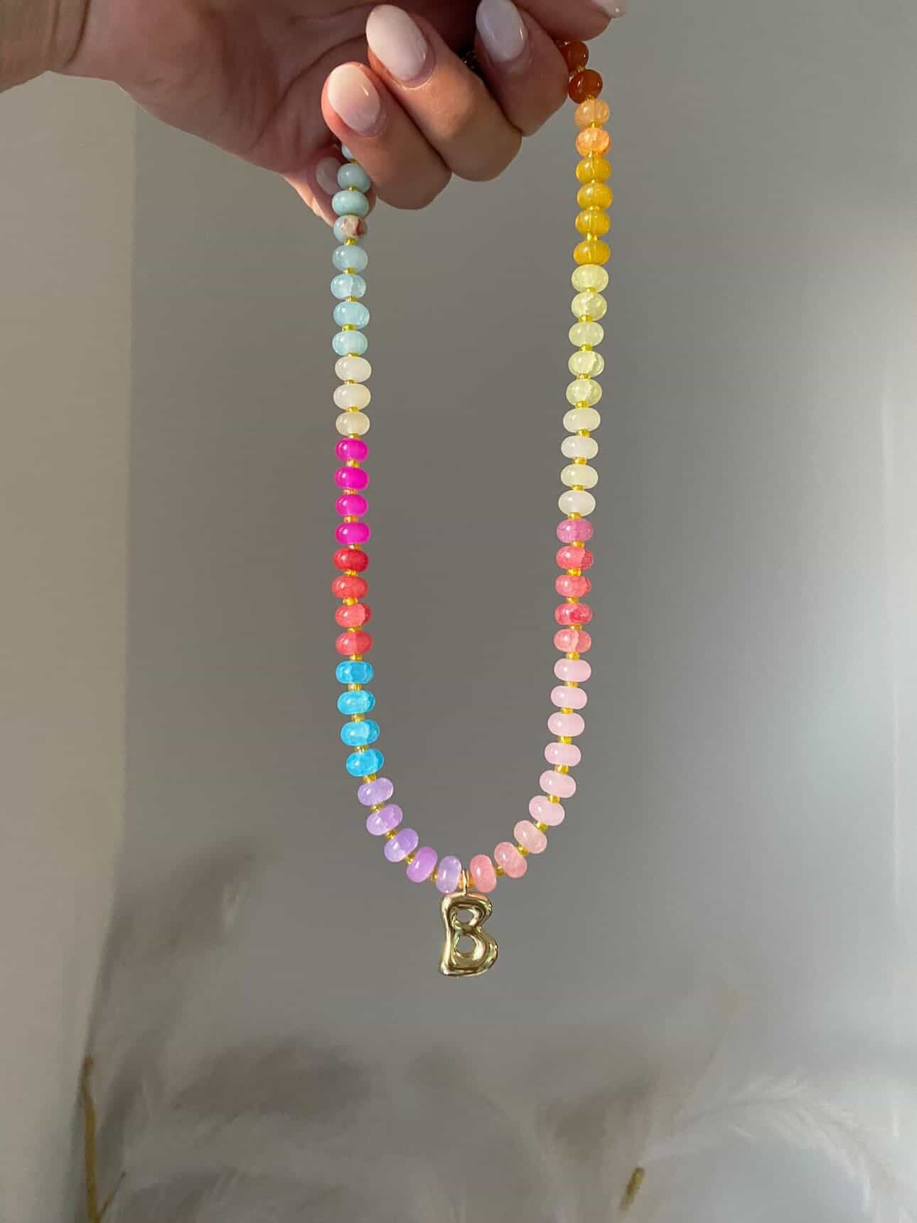 A multicoloured beaded necklace with tones of pale blue, lilac, pinks, yellows, and corals, separated by spacer beads. A gold bubble initial B hanging from the centre
