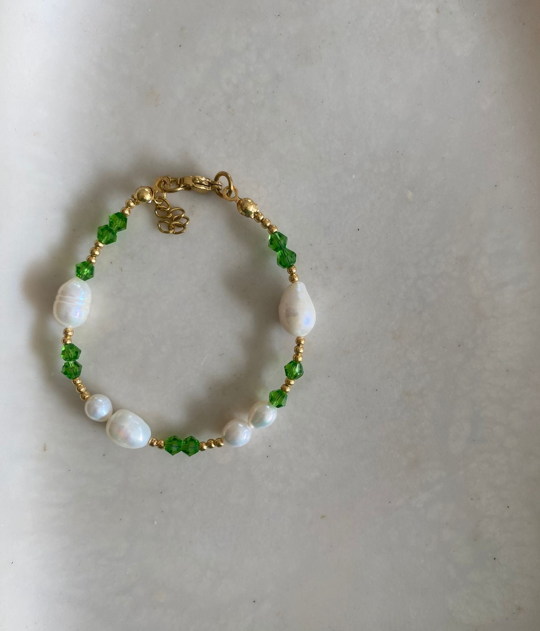 Bracelet with claw clasp, freshwater pearls, emerald green crystal beads, and gold seed beads, laid flat on a marble dish.