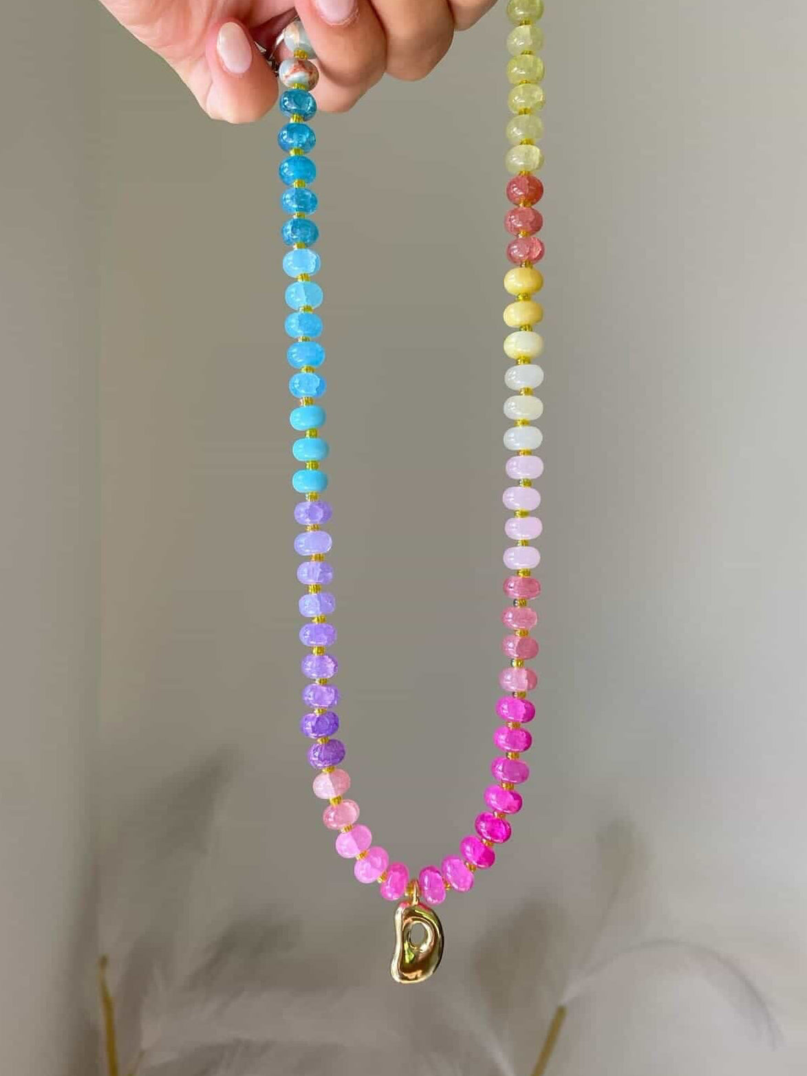A multicoloured beaded necklace with tones of blue, lilac, pinks, yellows, and corals, separated by spacer beads. A gold bubble initial D hanging from the centre.