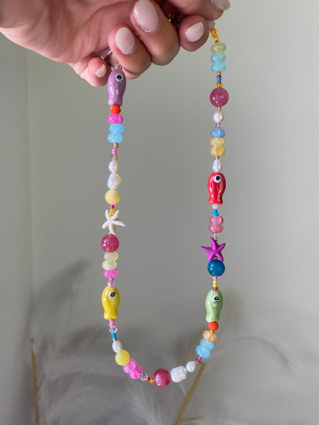 Beaded necklace featuring colourful gemstone beads, freshwater pearls, rainbow seed beads, and ceramic fish charms in purple, red, yellow, and green, with starfish accents