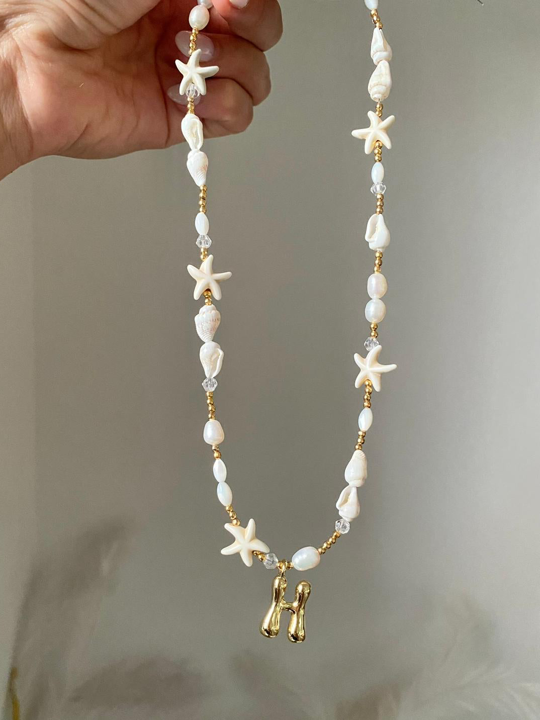 Beaded necklace featuring freshwater pearls, natural white shell beads, starfish charms, gold seed beads, and a gold bubble letter H initial pendant, held up against a soft grey background.