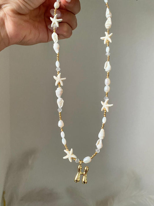 Beaded necklace featuring freshwater pearls, natural white shell beads, starfish charms, gold seed beads, and a gold bubble letter H initial pendant, held up against a soft grey background.