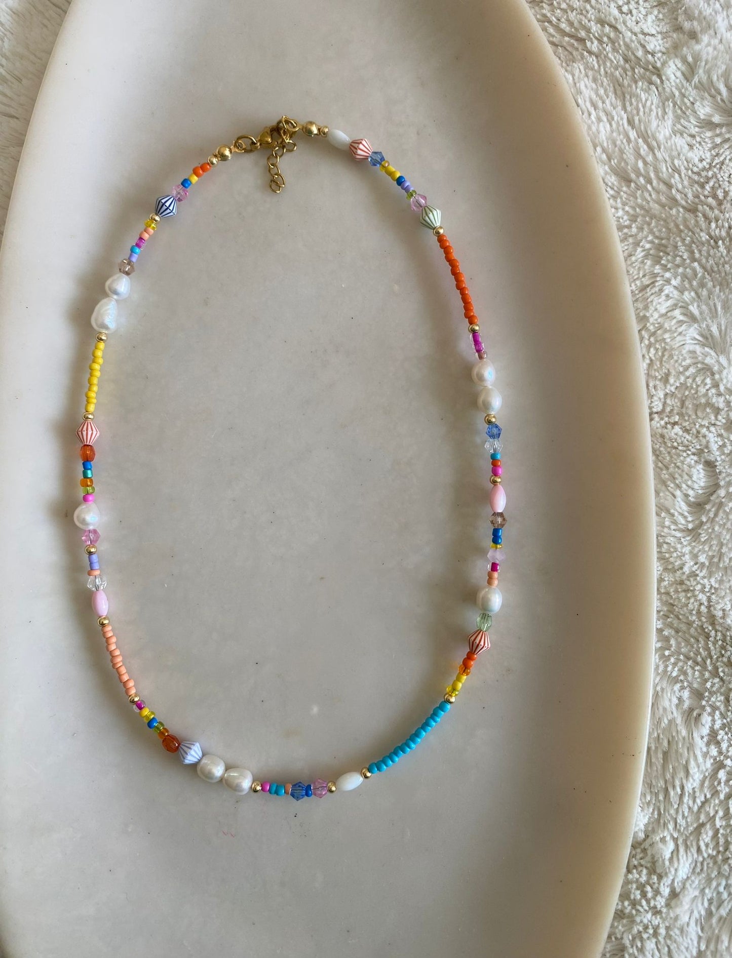 Beaded necklace with multicolour seed beads, freshwater pearls, crystal spacers, gold seed beads, and striped bicone charms laid flat on a marble dish