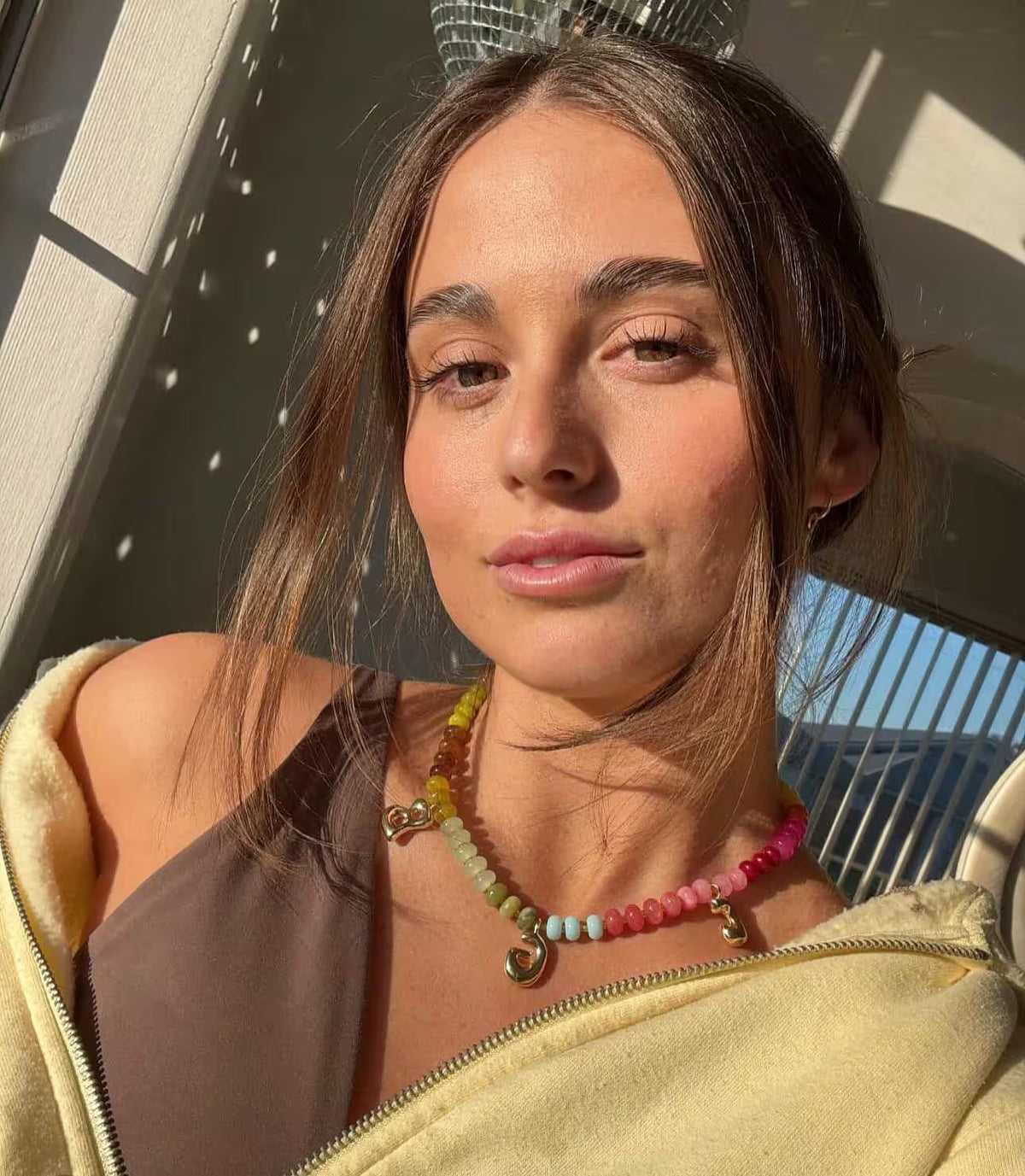 Selfie from a customer wearing a customised multicoloured gemstone necklace with gold bubble initial charms, styled with a yellow hoodie and brown top