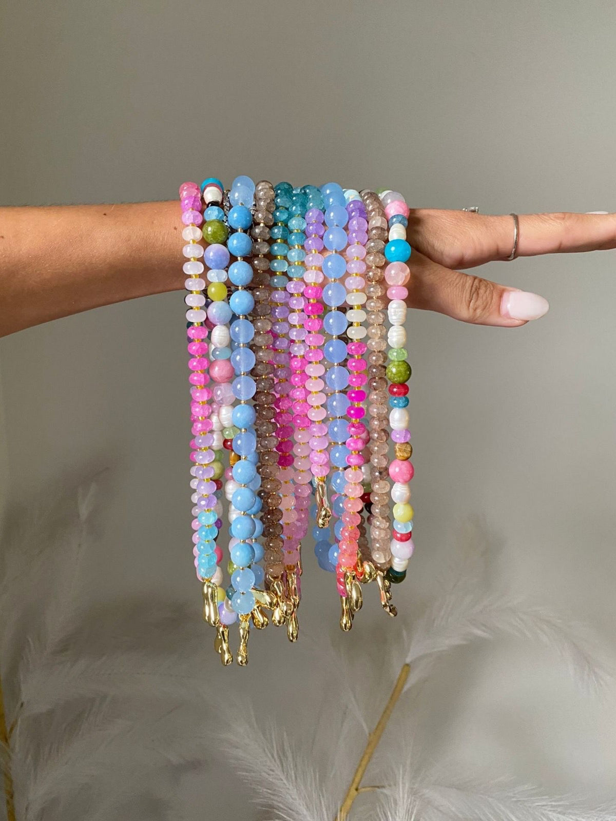 A bundle of 10 colourful beaded necklaces with gold initial charms, hanging over Ele's wrist and dangling down. 
